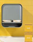 joni Model A open dispenser for period care pads and tampons hanging on a yellow brick wall with boxes of joni tampons