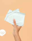 Hand holding period pads with a 'Donate Now' badge for Karma Donation Pack to support those in need.