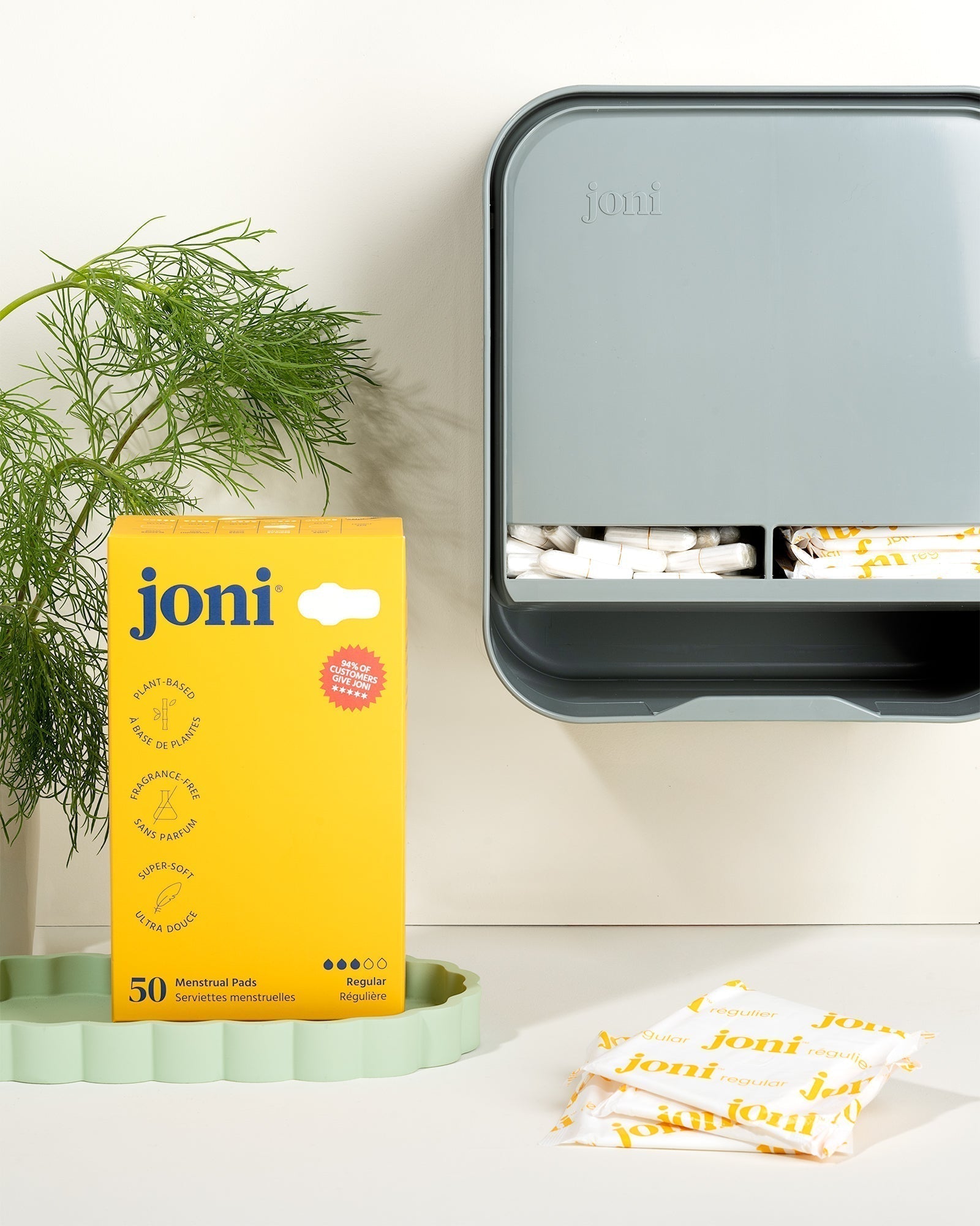 A joni model a open period care dispenser hangs on a white wall with a box of joni regular pads on the countertop