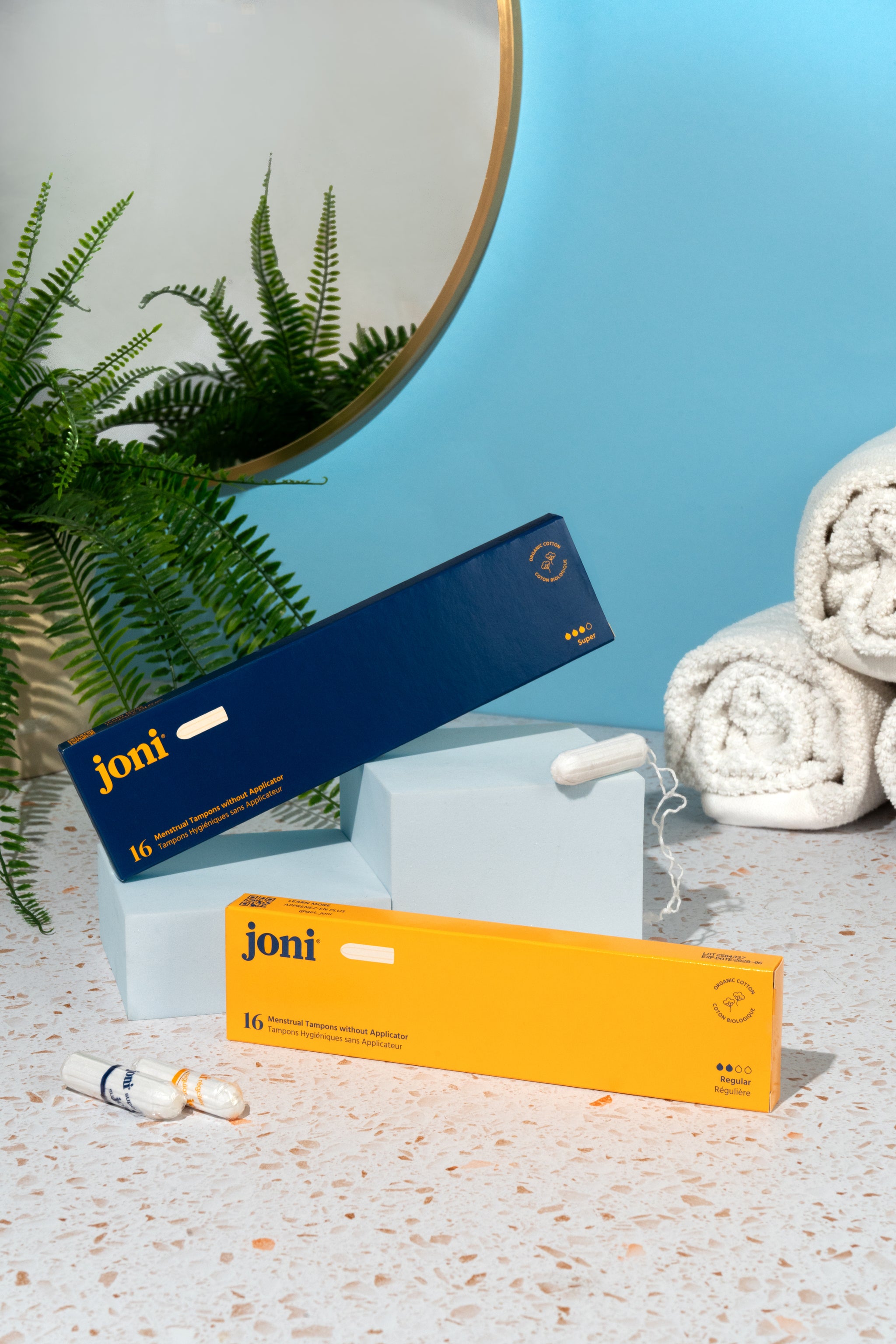 Two boxes of joni organic cotton tampons without applicators on a surface with a plant and towel in the background.