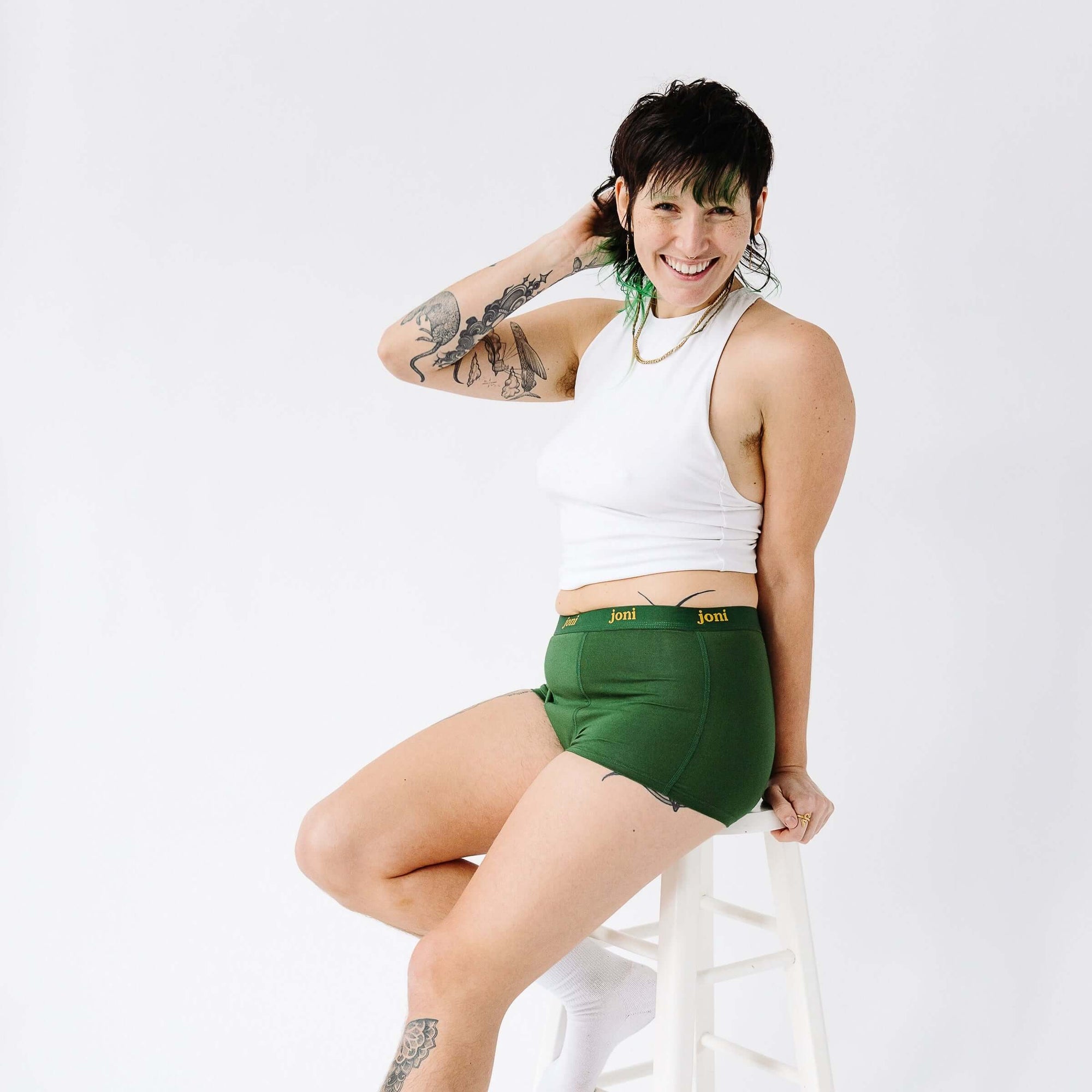 Person in joni green period underwear sits on a stool wearing a white tank top