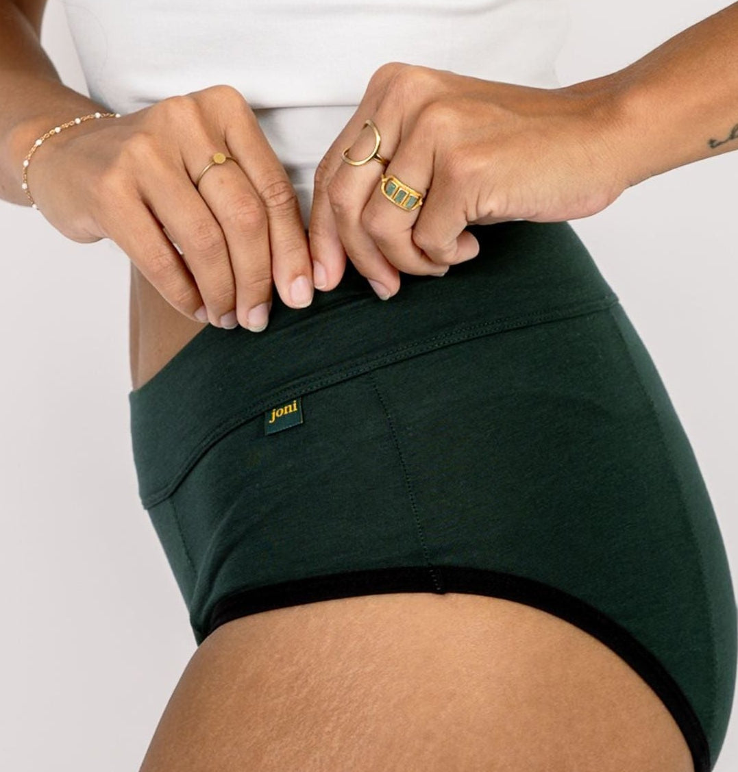 A close-up of a model wearing dark forest green joni period underwear with a small yellow and green logo tab on the front left pocket, paired with a white tank top against a light gray background.