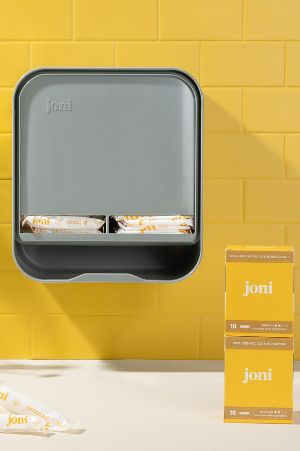joni Model A open dispenser for period care pads and tampons hanging on a yellow brick wall with boxes of joni tampons