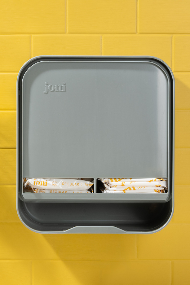 joni Model A open period care dispenser on a yellow brick wall