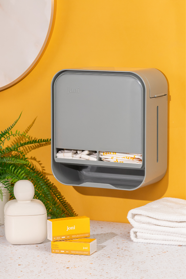 Wall-mounted joni Model A period pad and tampon dispenser in a yellow washroom with a plant